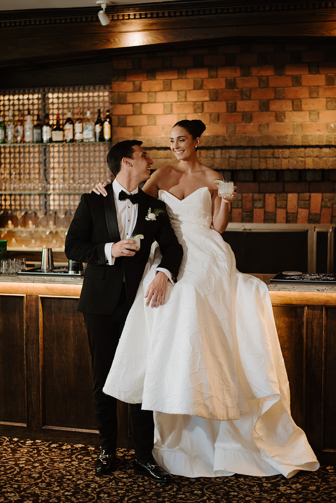 Bride sitting on the bar counter smiling at the groom as they hold cocktails with lime wedges.
